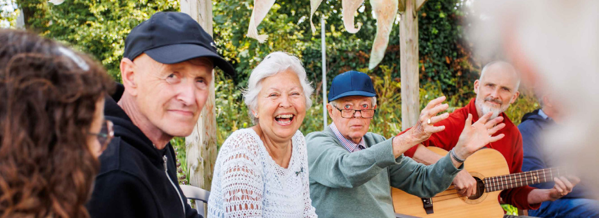 A row of people sitting outdoors. One has a guitar, one is dancing in his chair and two are looking to camera.