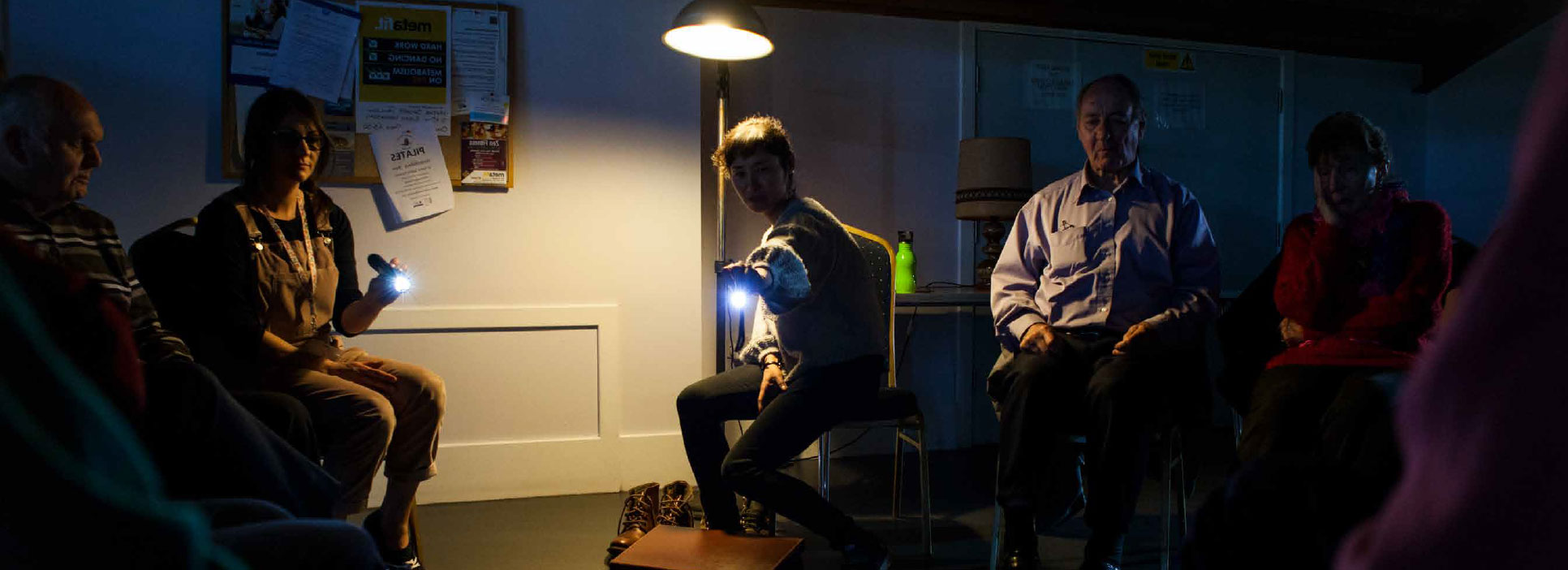 A room lit by torches, with two rows of people facing each other, looking at a performer who is sitting down in the top left corner.