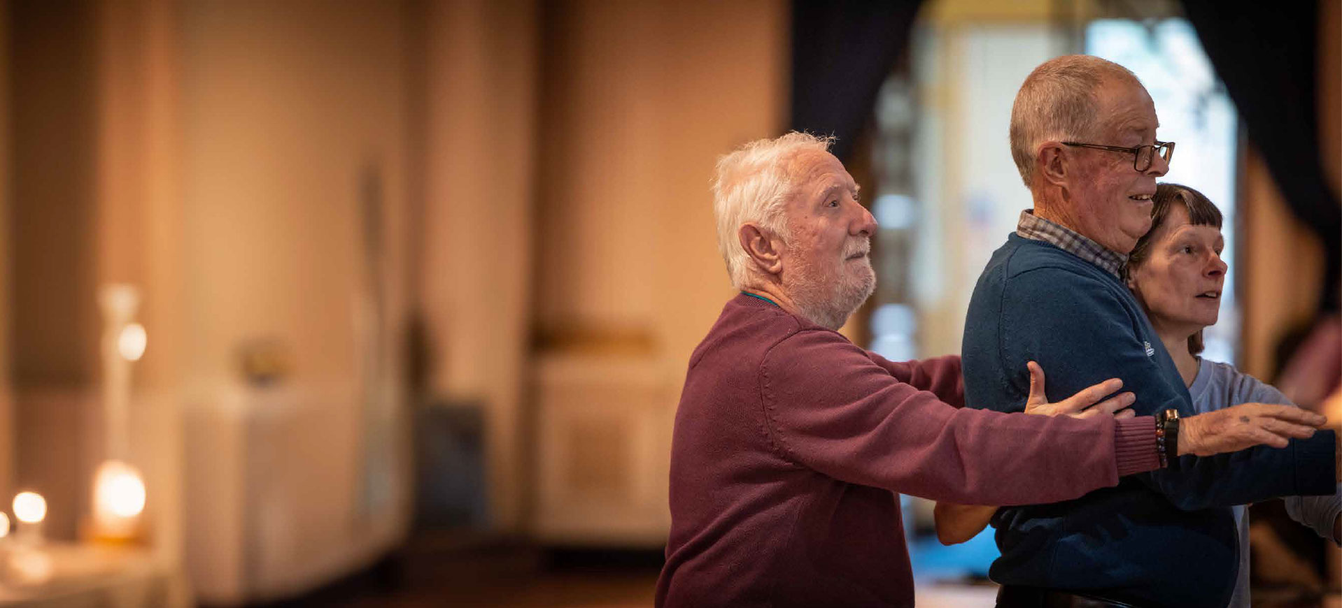Two male participants are stood in front of a younger woman. They are all facing right, with their arms raised out wide in front of them, at shoulder height.