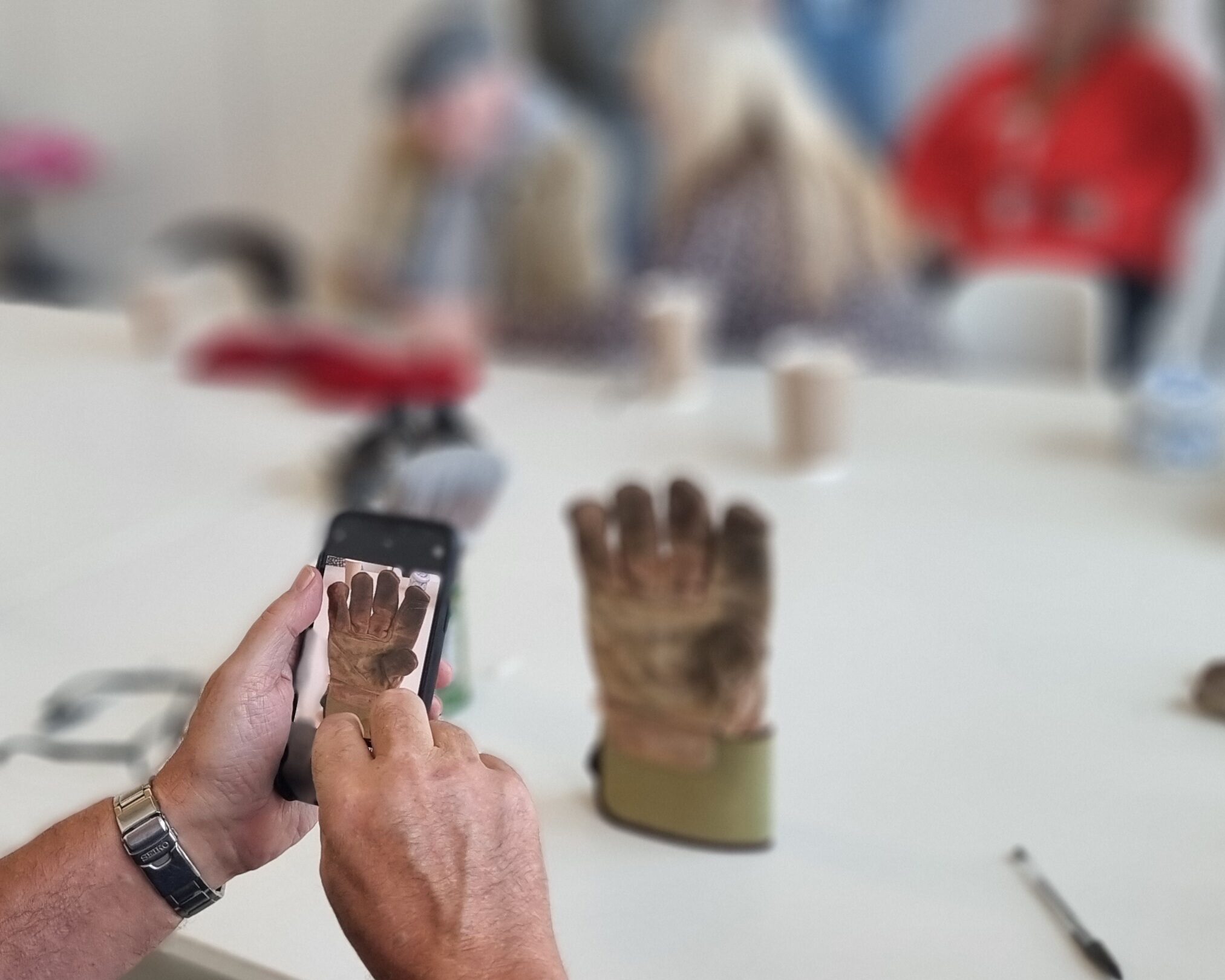 a man is using his phone to take an abstract photograph of a gardening glove stood up on a white table.