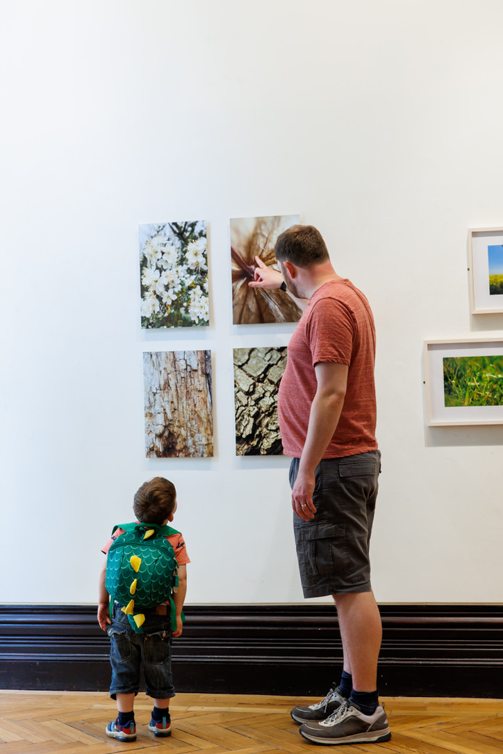 A man in a red t shirt and green shorts stands with a boy with a green dinosaur backpack to look at close up photographs of the natural world hung on the wall of an exhibition in a gallery space.