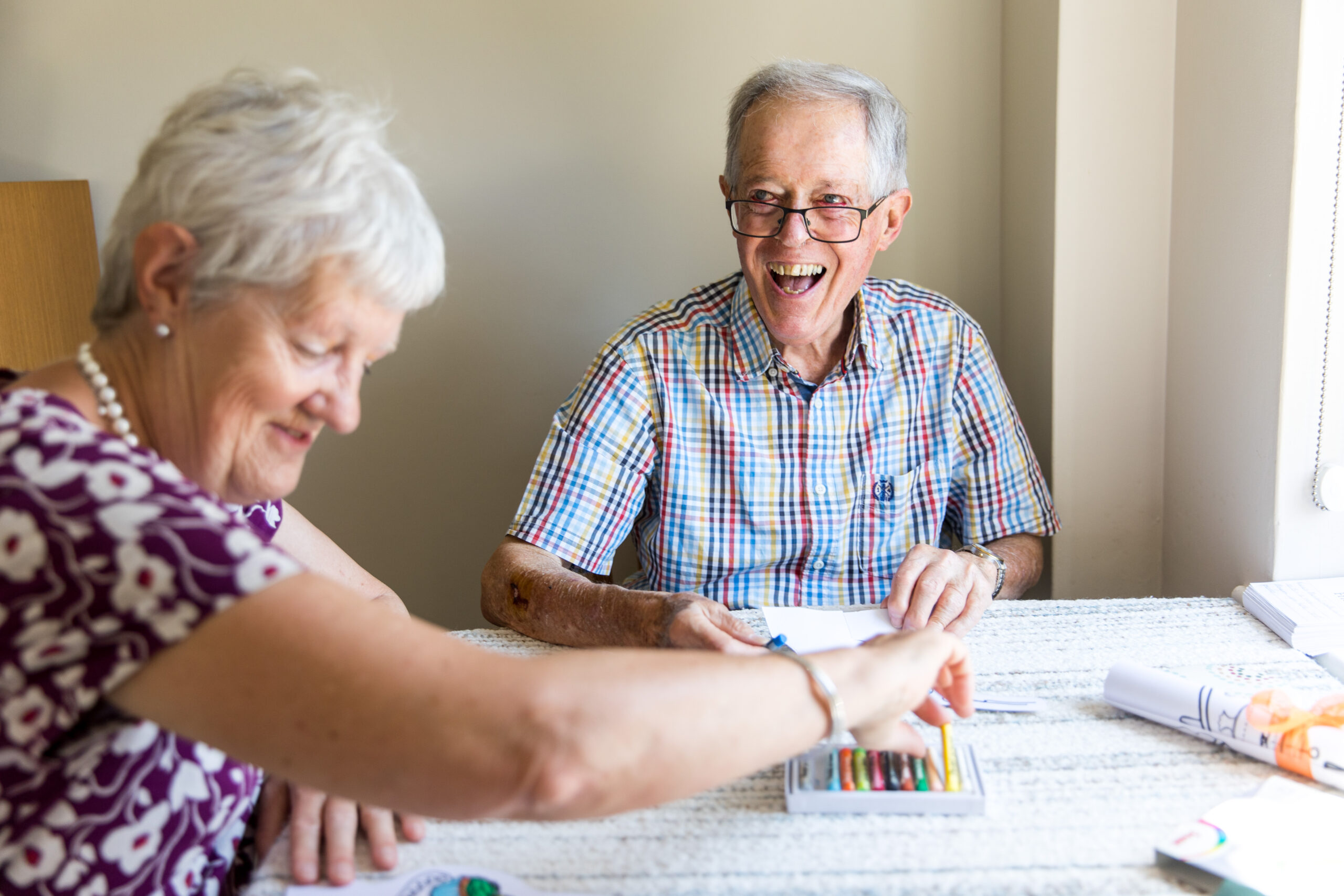 A man and a woman are sitting at a table, laughing. They are using oil pastels to make patterns on a colouring activity sheet.