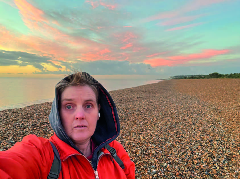 A woman in a red jacket with her hood up stands on a shingle beach with a colourful sky behind her. She is taking a self-portrait.
