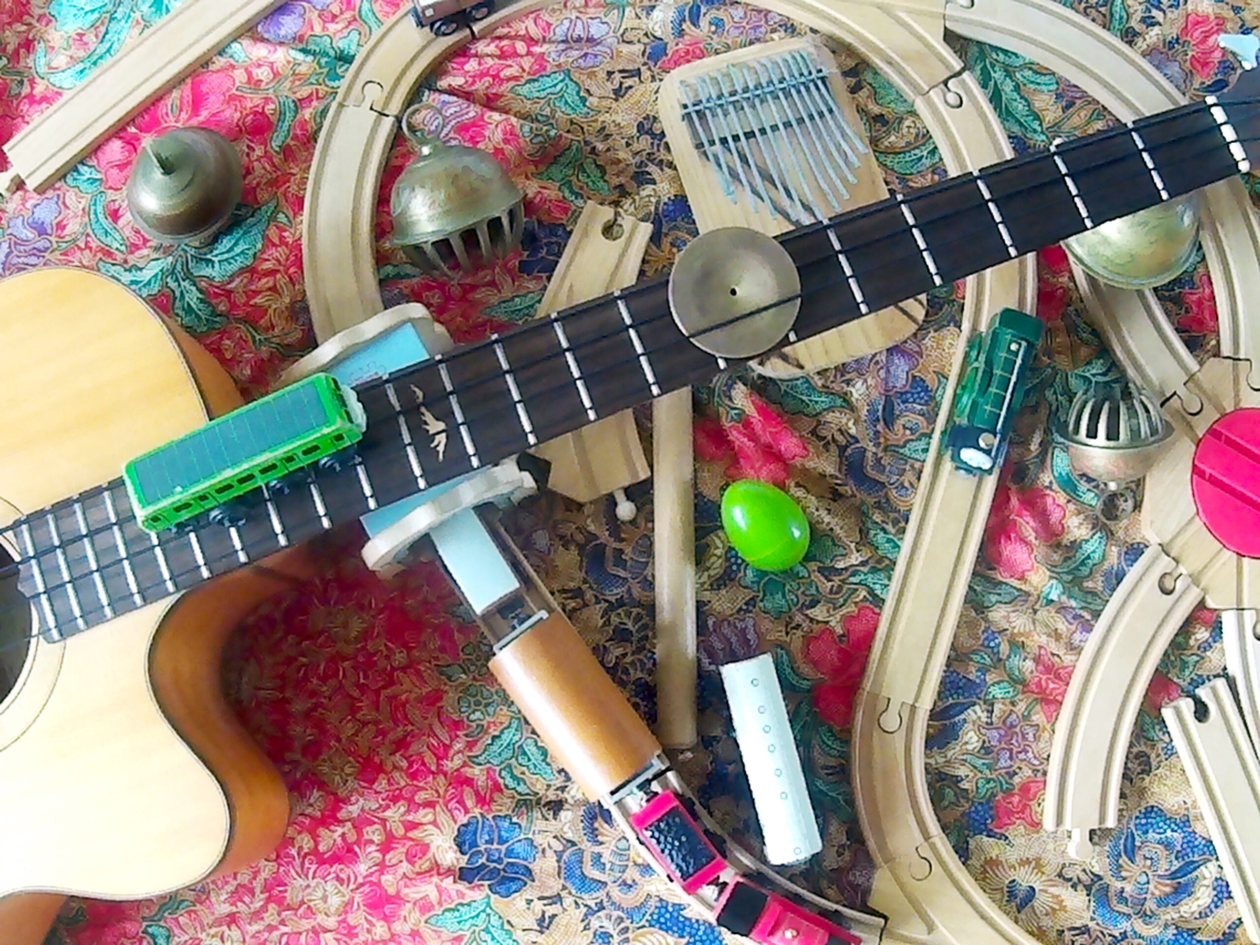 The neck of a guitar lies across a toy train set made of wood. One of the toy trains is sitting on top of the guitar.
