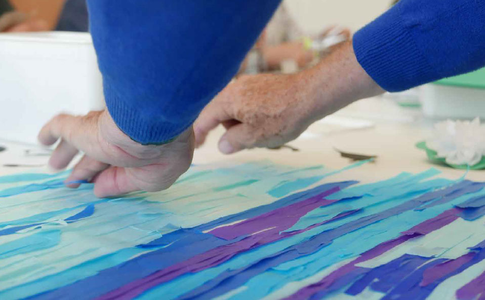 A close up of a tissue paper collage in several tones of blue. Someone is reaching out to place more tissue paper down.