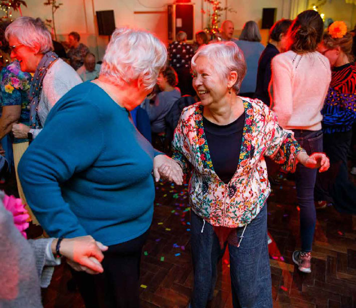 Three women are holding hands, two are facing each other, dancing and smiling. They are in a big room filled with other people dancing and talking. The lights in the room offer a warm glow.