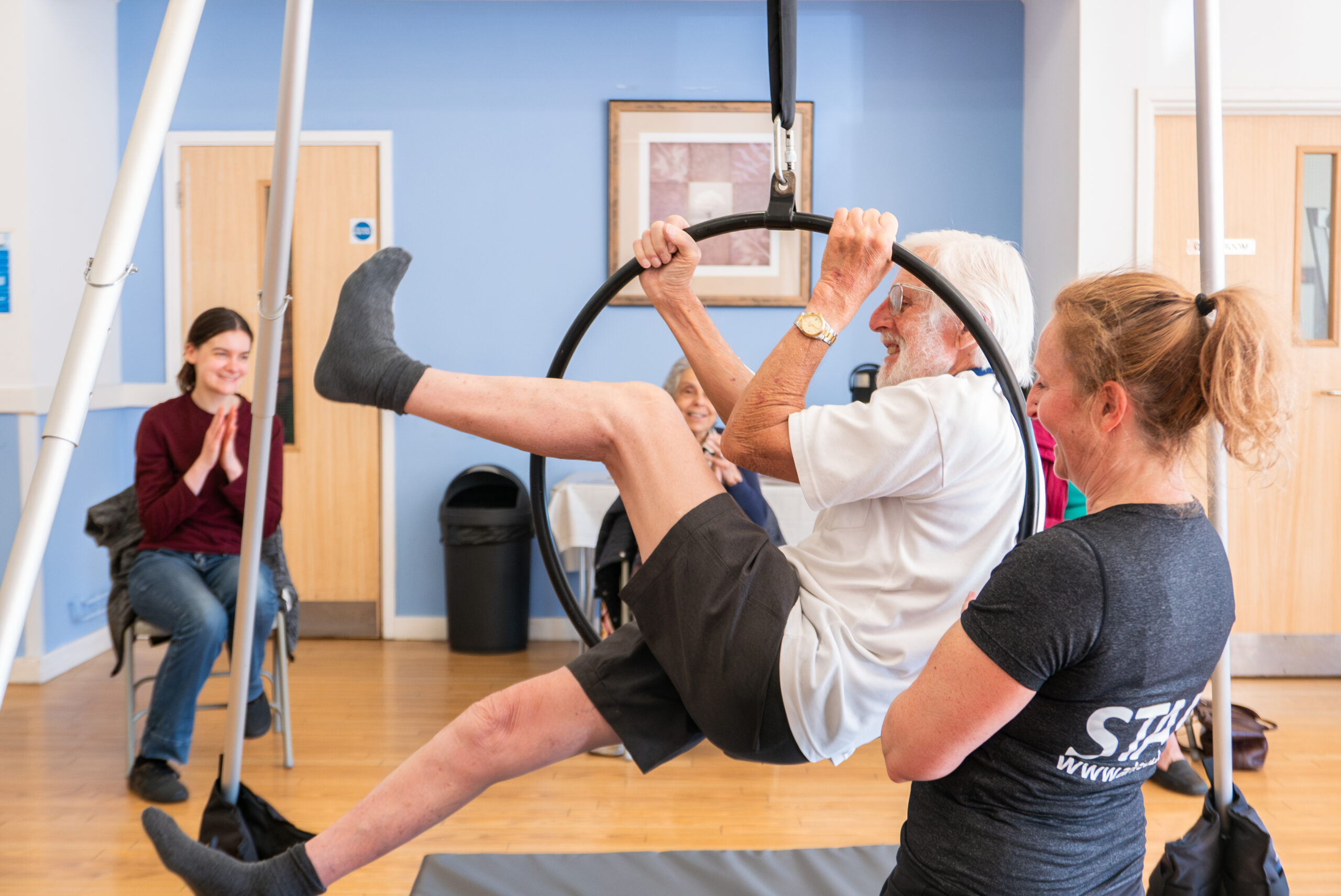 A woman supports an older man who is suspended on an aerial hoop, above the ground. He is facing left, using his arms to hold himself in the hoop as his legs help to balance him above the ground.