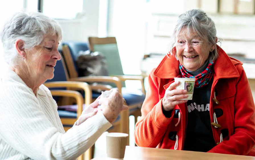 Two women are sitting at the corner of a table, each with a hot drink. They are smiling.