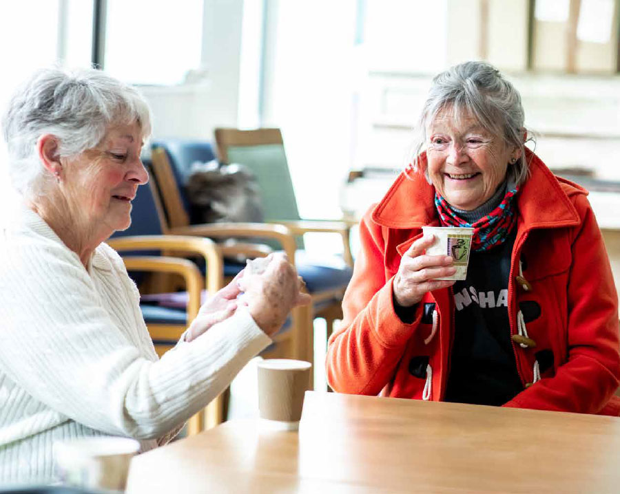 Two women are sitting at the corner of a table, each with a hot drink. They are smiling.