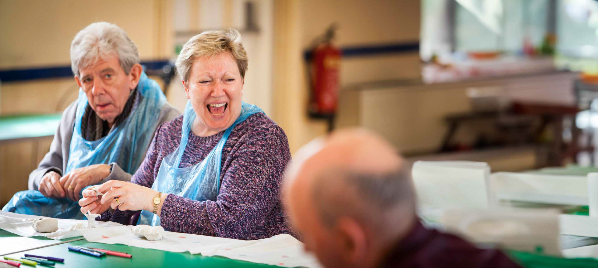 A man and woman wearing aprons sit at a table making items from clay. The woman is laughing with someone not in the picture.