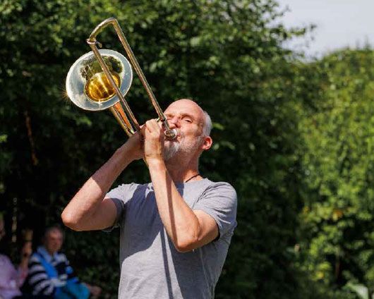 a man plays a trumpet outdoors in front of green trees on a sunny day