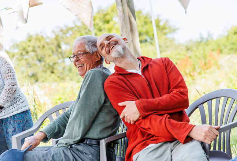 Two men are sitting outdoors on garden furniture. They are leaning on each others' shoulders and smiling.