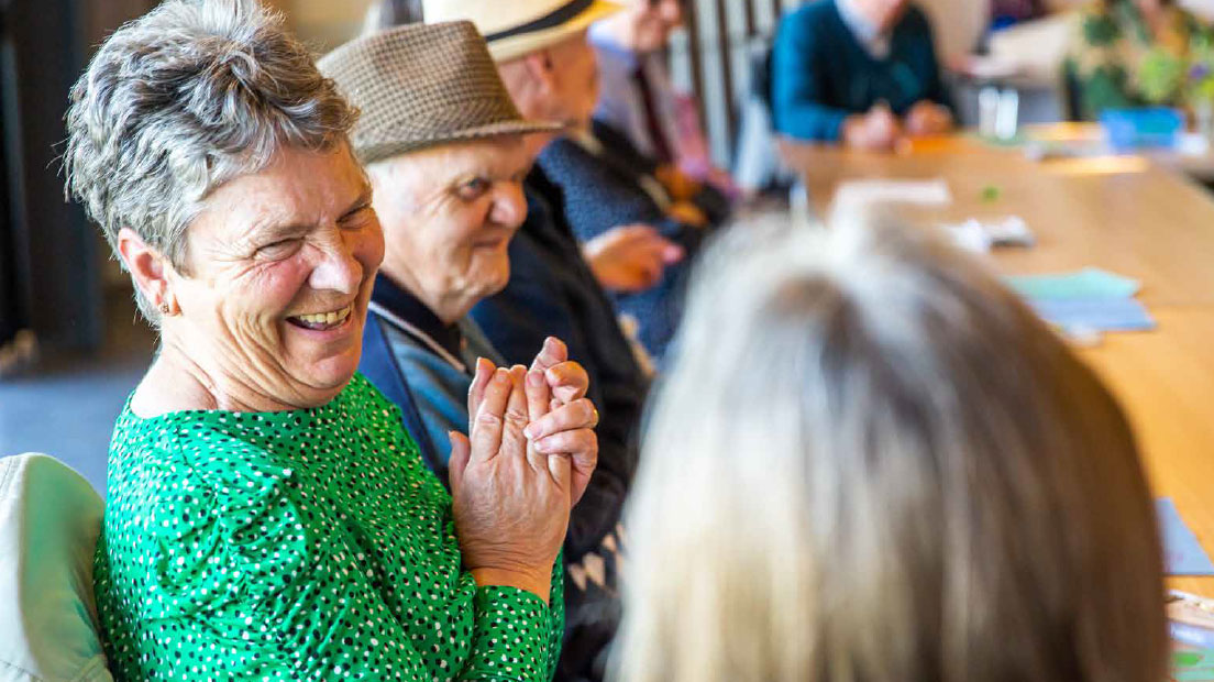 A group of people are sitting around a table in a bright, airy room. In the foreground, a man and a woman are smiling and looking to people sitting to the right of the photo.