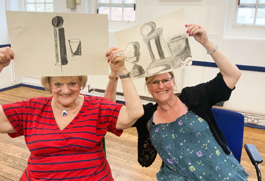 impact-slide-1 Two women are sitting down, smiling at the camera and holding up charcoal drawings above their heads.