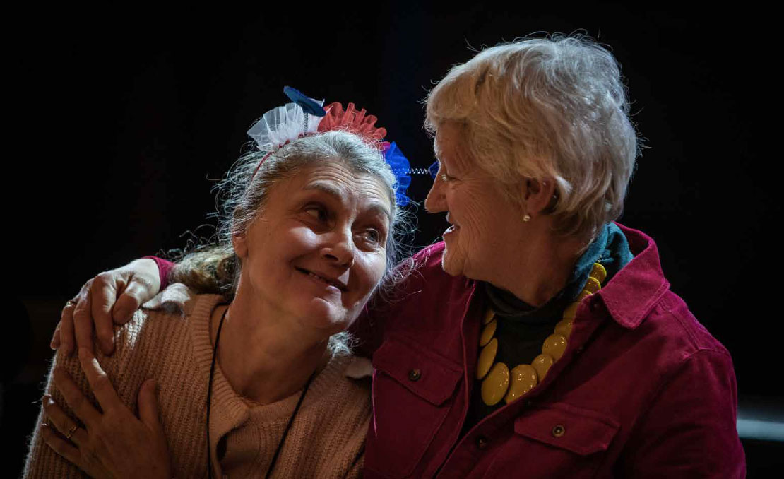 impact-slide-2 Two women are sitting in front of a dark curtain lit by theatre lights. One has her arm around the other, and they are looking at each other, smiling.