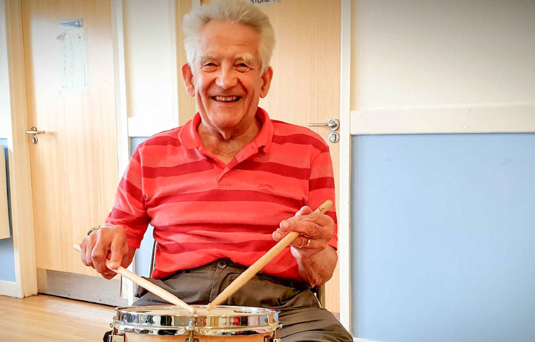 impact-slide-3 A man is seated behind a drum on a stand, holding a drumstick in each hand and smiling.