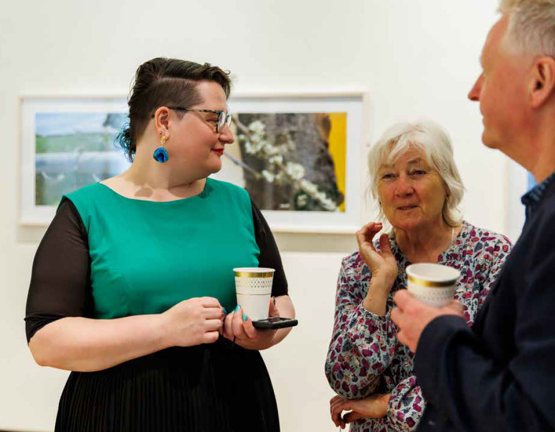 Three people stand in an art gallery holding drinks and talking.