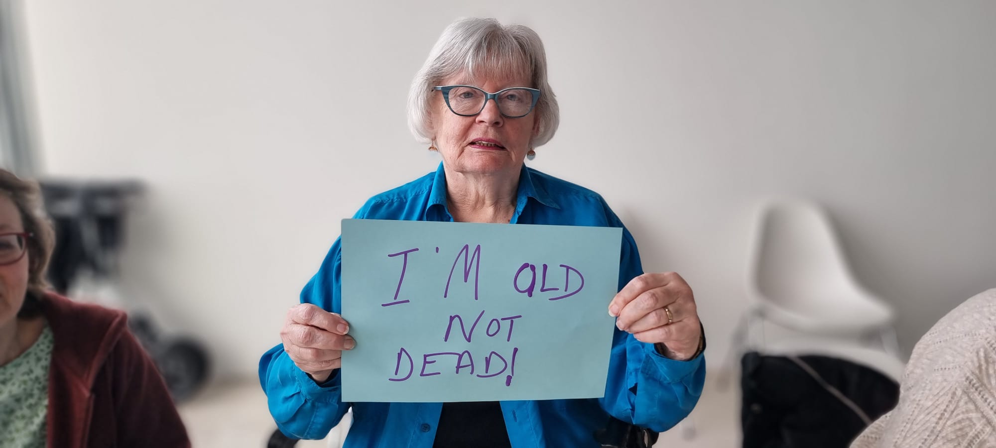 Advocacy-slide-3 A woman wearing glasses holds up a handwritten sign which reads I'm old but I'm not dead!