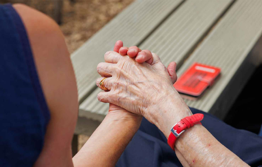 volunteer-3 Two people are sitting outside near a picnic bench. The photo is taken from behind, and shows them holding hands.