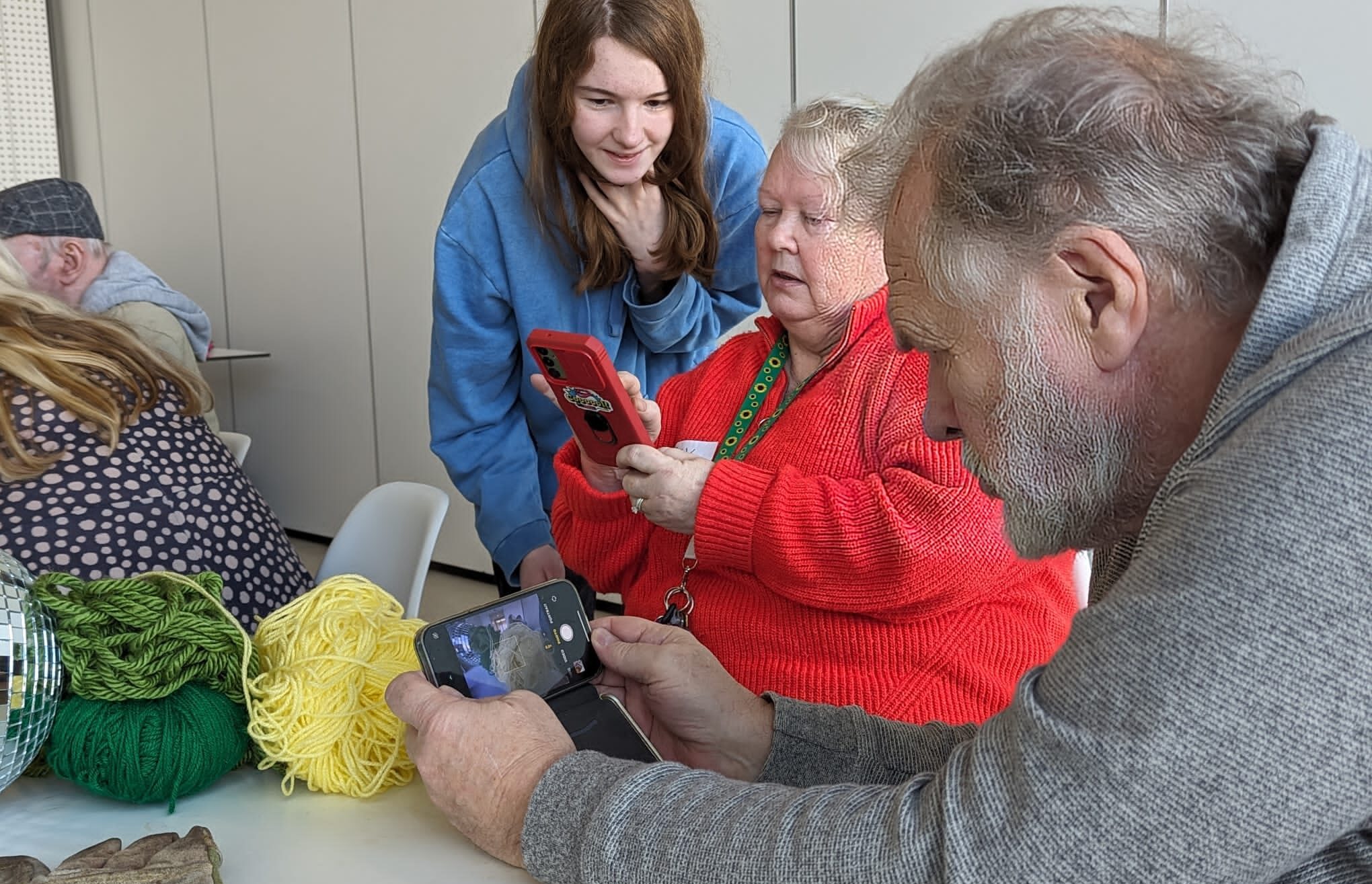 volunteer-read-more-2 A volunteer stands behind two people sat at a table. The volunteer is helping one with her phone, as they take photographs.