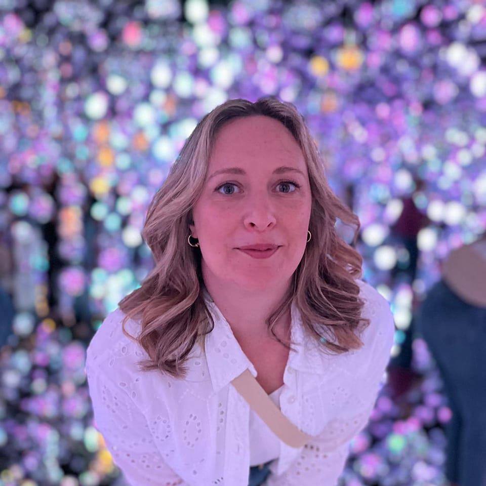 BeckyVincer a white woman with mid-length blonde hair stands in front of a multi-coloured twinkly background. She is looking up to the camera.