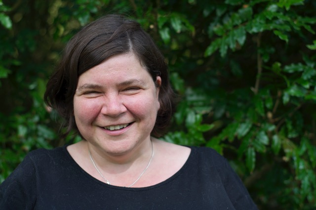 A white woman is stood in front of a leafy tree, smiling to camera