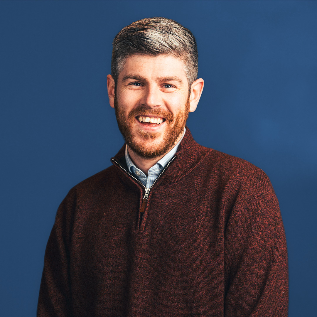 A white man stands in front of a blue background, smiling to camera