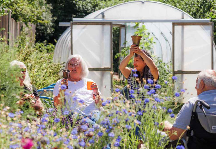 Three women and a man are sitting in a garden, surrounded by flowers, with a poly tunnel behind them. Two of the women are ringing bells.