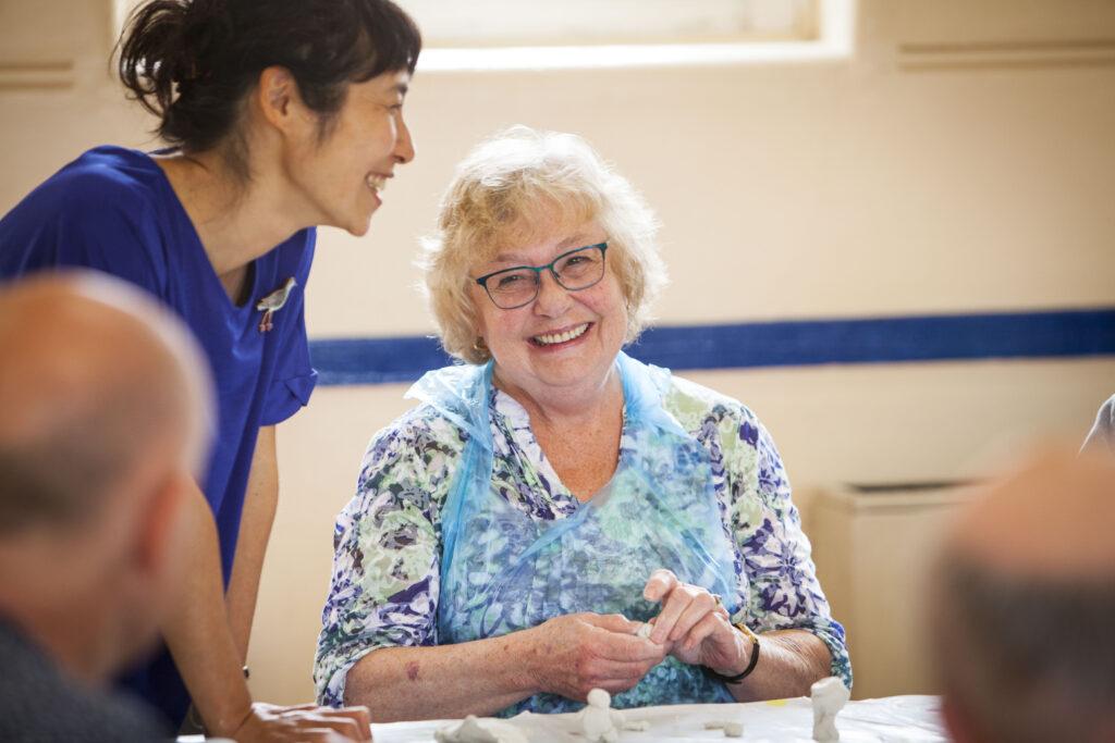 A woman with grey curly hair, wearing glasses and a blue apron over a floral top, is smiling at the camera while making with her hands. There is a table in front and a wall with a window behind. A woman with black hair and a blue top is smiling next to her, with the backs of two men's heads visible in the foreground.