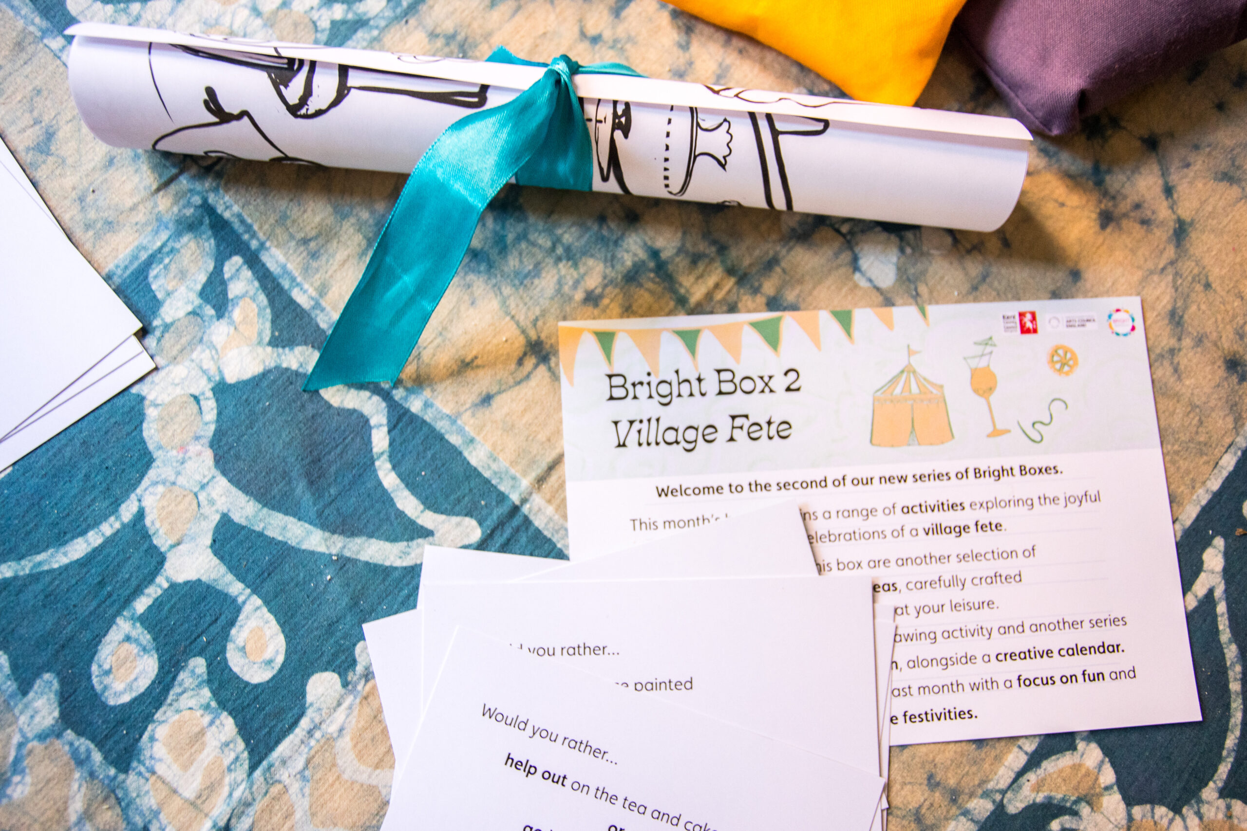 A selection of printed artistic resources, including some cards with a 'Would you rather' game and a rolled up colouring sheet fastened with a ribbon, are scattered across a kitchen table. The table is covered with a table cloth.