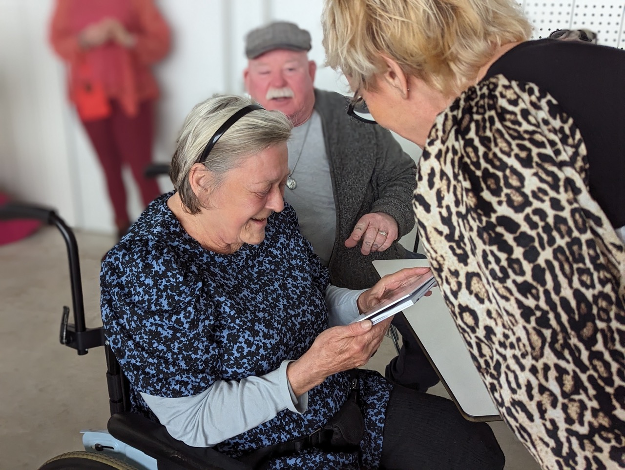 A grey-haired wheelchair-using woman in a blue patterned t shirt receives a book from a blonde-haired woman in leopard print. A woman dressed in red stands in the background behind a moustachioed man in a flat cap.