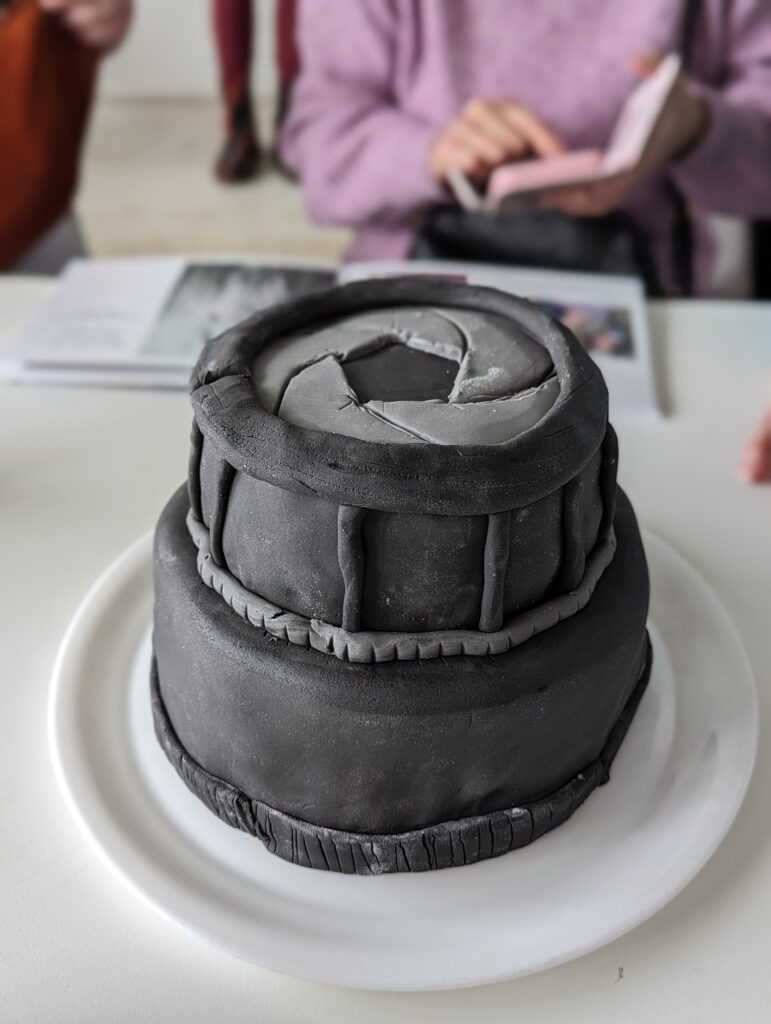 A plate with a tiered cake in the shape of an SLR camera with black icing and grey details.