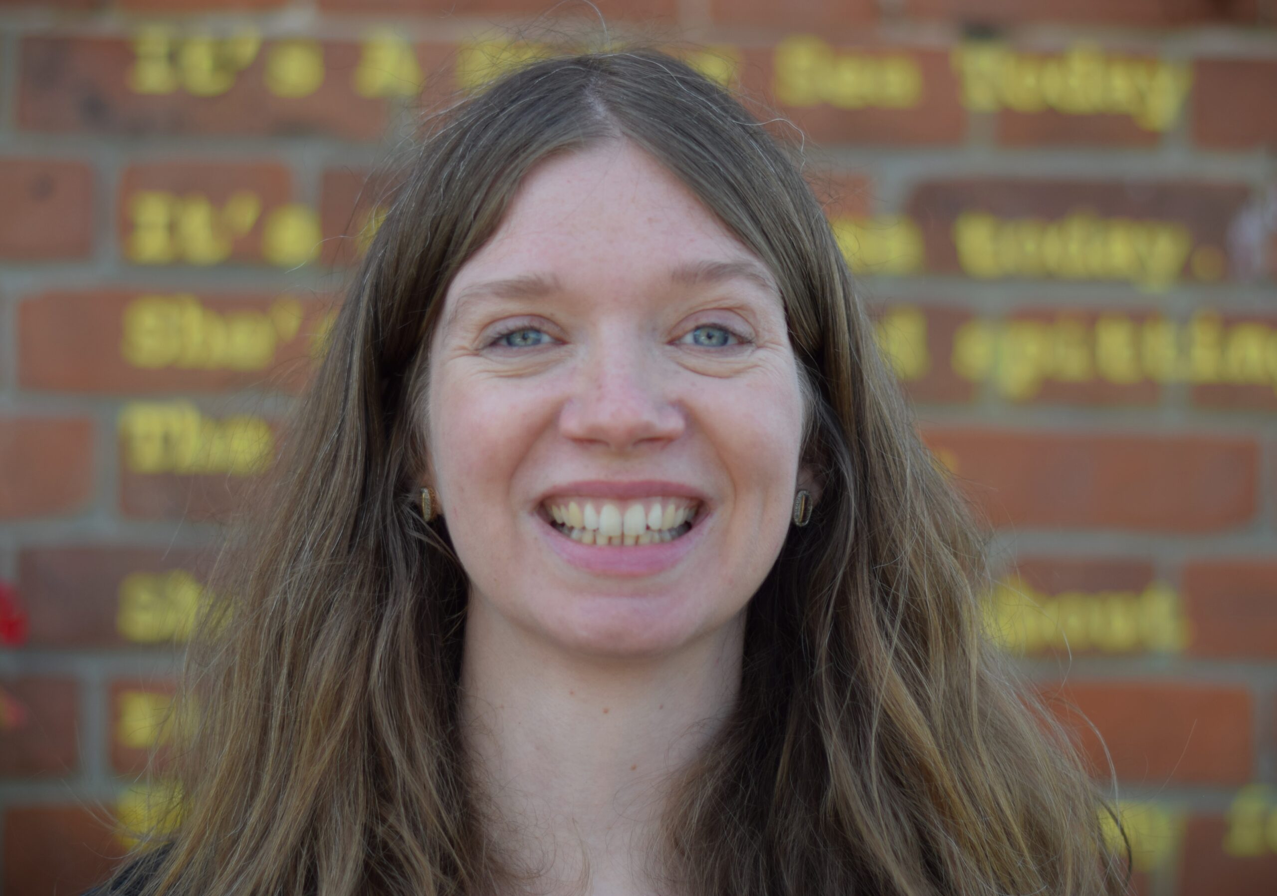 A white woman with long hair smiles and looks to camera in front of a red-brick background with some yellow text sprayed on that is not in focus
