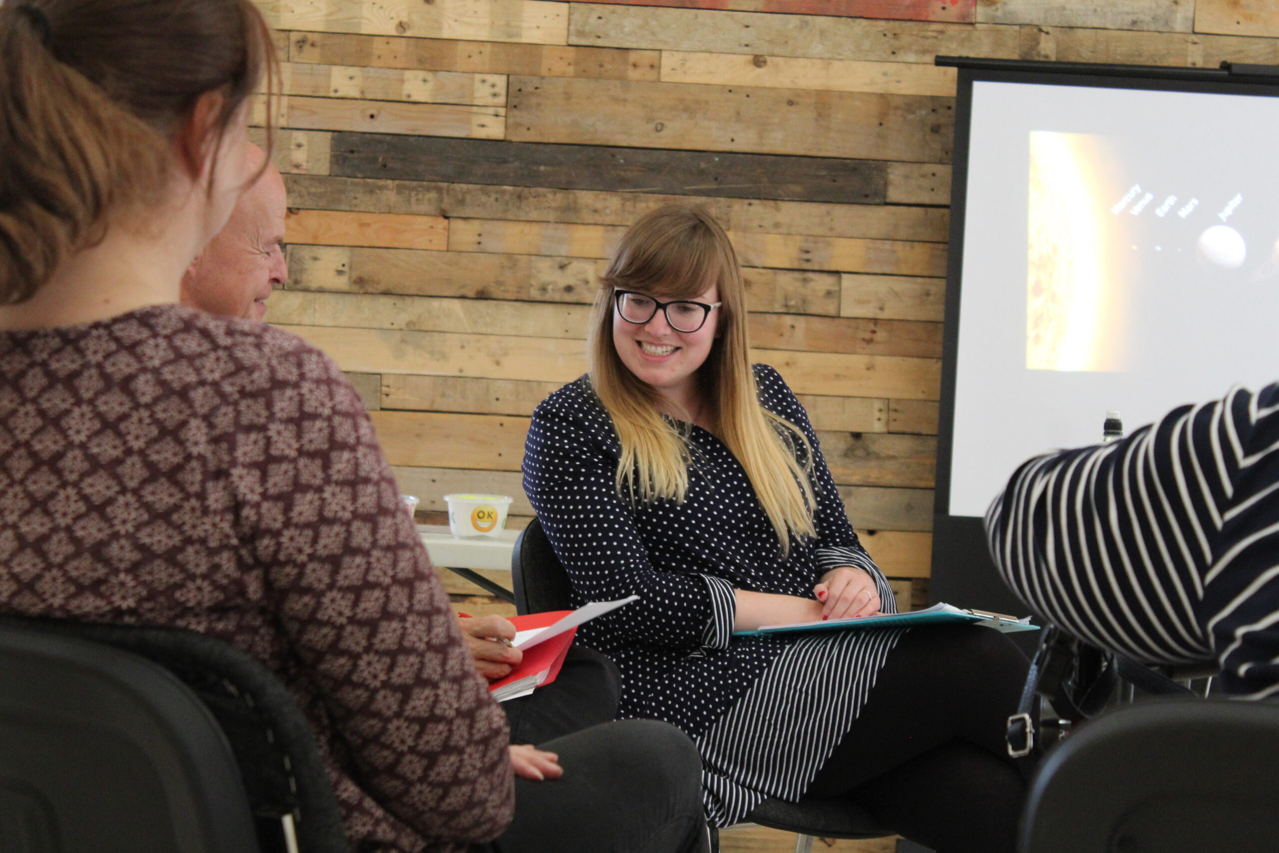 A group of people are sat in a bright room. They have training packs in their lap and a woman with glasses is smiling, looking across