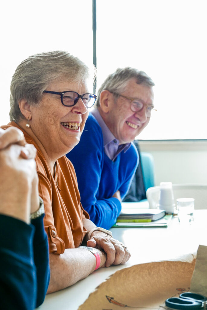 Three people are sitting beside each other. Only the first person's clasped hands are visible. Next to them are a woman in orange and a man in blue, both with short grey hair, smiling across the table. On the table are sriting materials and cups. Behind them is a screen.