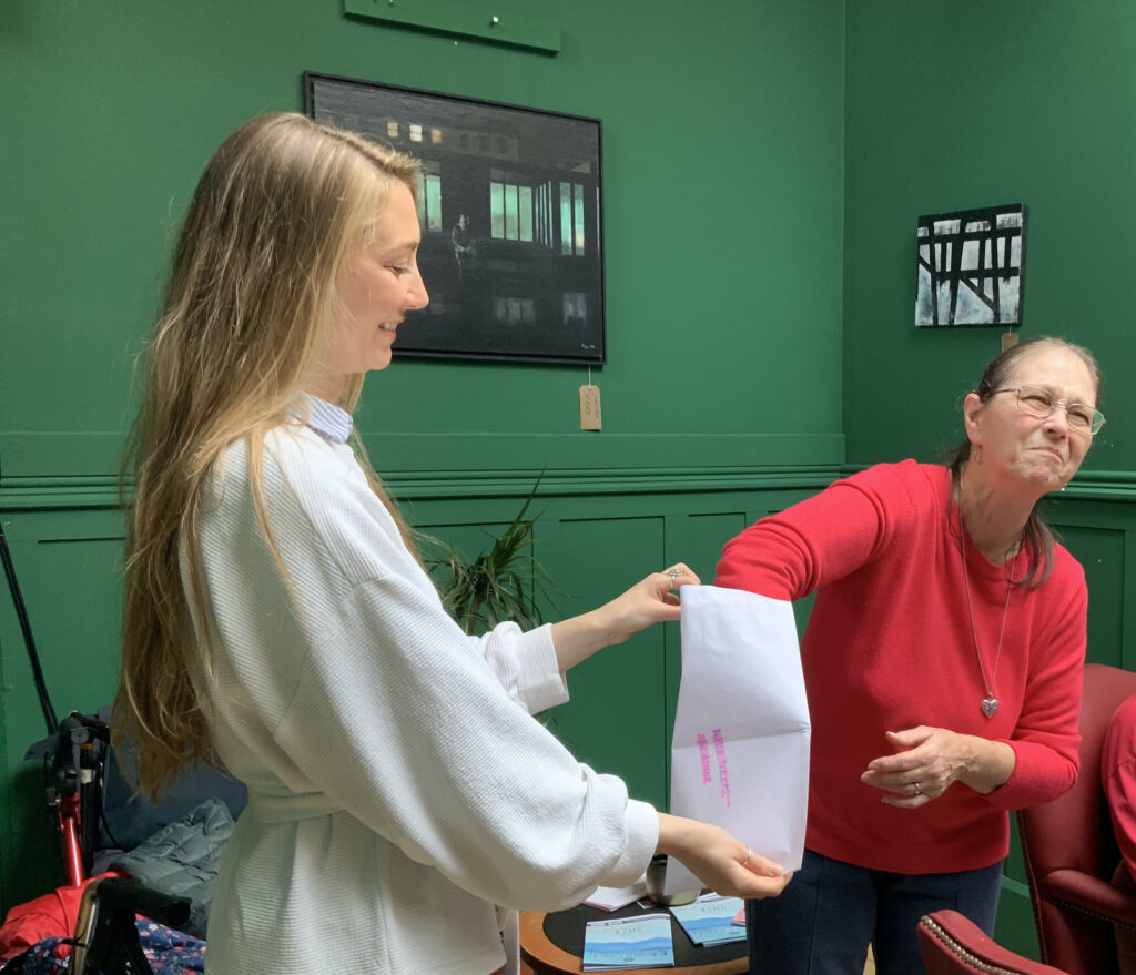 two women stand in front of a green wall. One in white holds the raffle envelope whilst one wearing red pulls the winning number