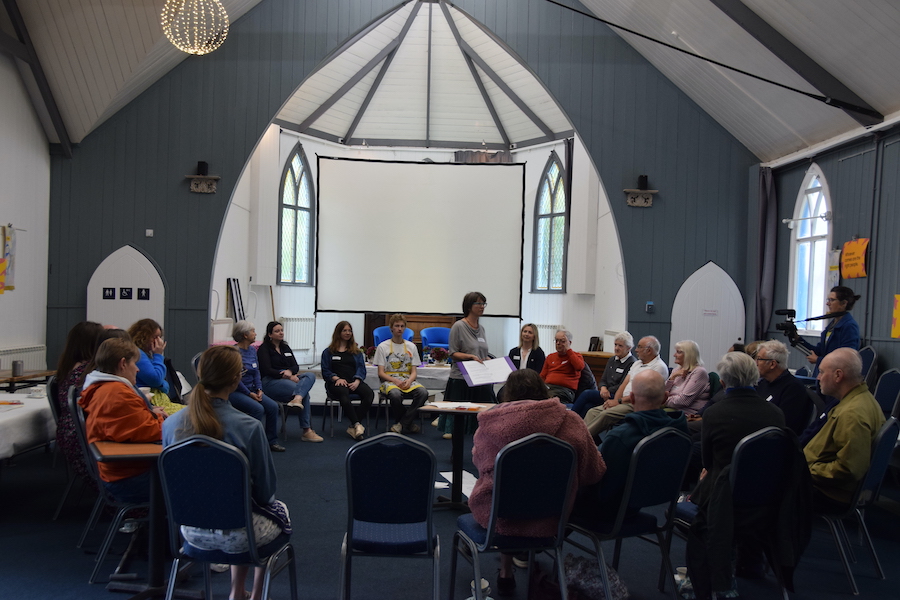 Open Forum 10 A wide shot of a group of people sat in a circle in a bright room. A woman is stood in the middle of the circle, talking to the group.