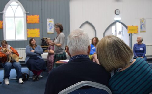 A group of people are sat in a circle. there is a woman in the middle who appears to be mid-conversation.
