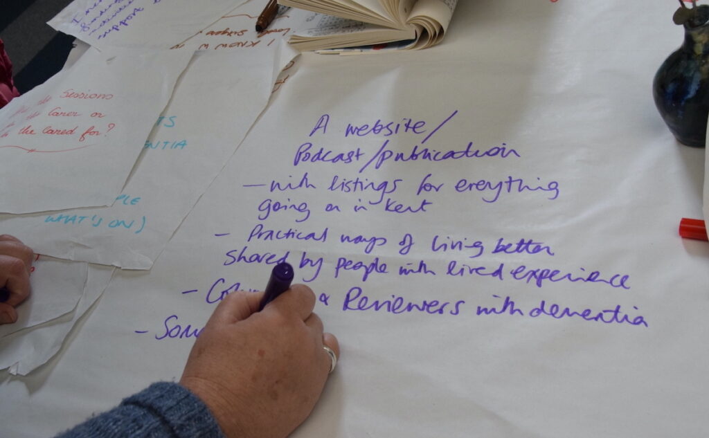 A photo of someone writing notes in purple ink. The words read a website / podcast / publication with listings for everything going on in Kent practical ways of living better hared be people with lived experience columnists and reviewers with dementia The scribe is continuing to write more.