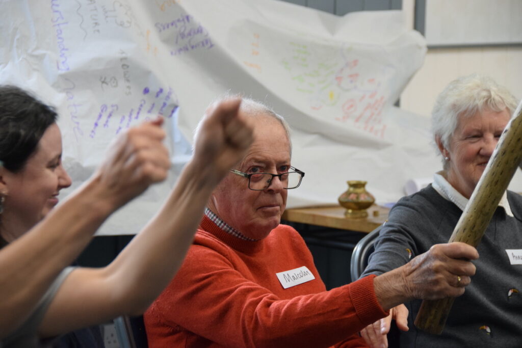 An older man in an orange jumper is holding a stick and looking to someone outside of the photo. either side of him are smiling women.