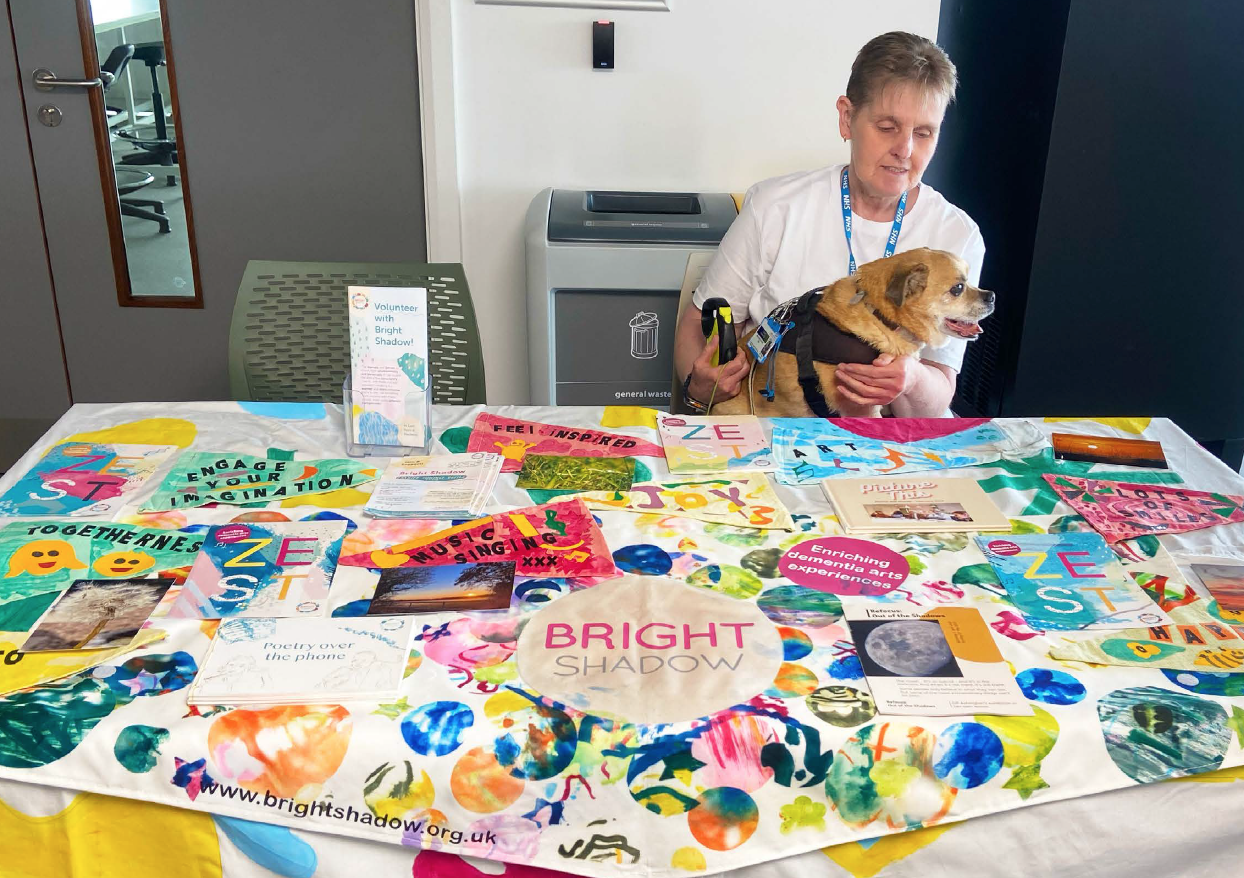 Gill and Rocky at CCCU conference A woman sits with a dog on her lap behind a table. The table has brightly coloured tablecloth and leaflets about Bright Shadow adorning it.
