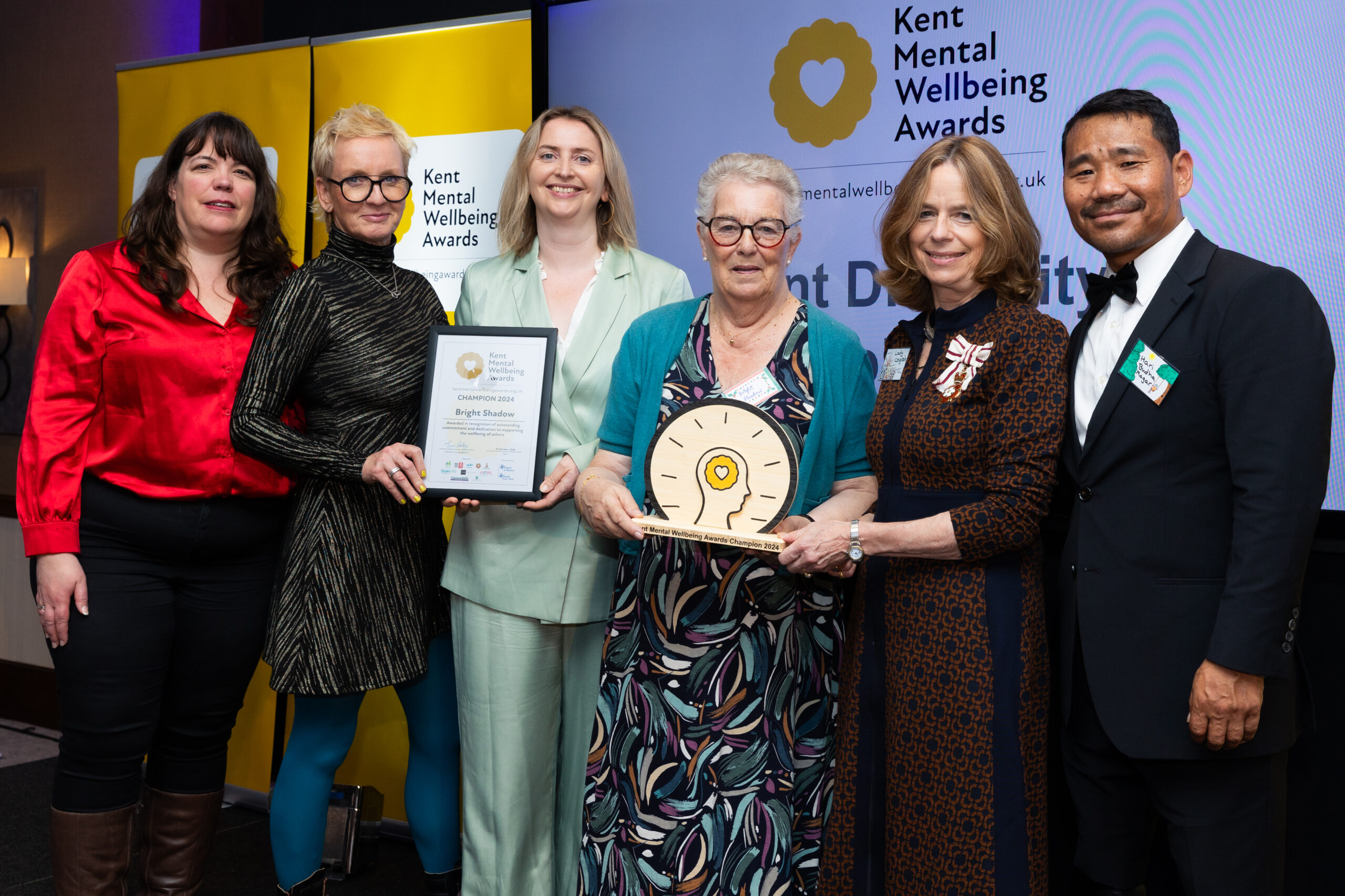 A group of people stand in front of the Kent Mental Wellbeing Awards banner, holding a champions certificate and award. They are smiling to camera.