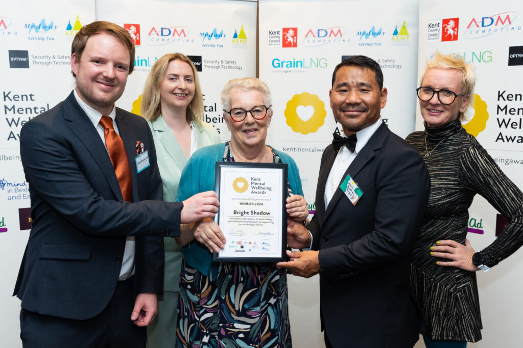 A group of people stand in front of the Kent Mental Wellbeing Awards banner, holding a champions certificate and award. They are smiling to camera.