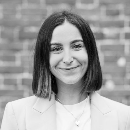 A white woman with brown hair smiles to camera. She is wearing a blazer, and stood in front of a brick wall