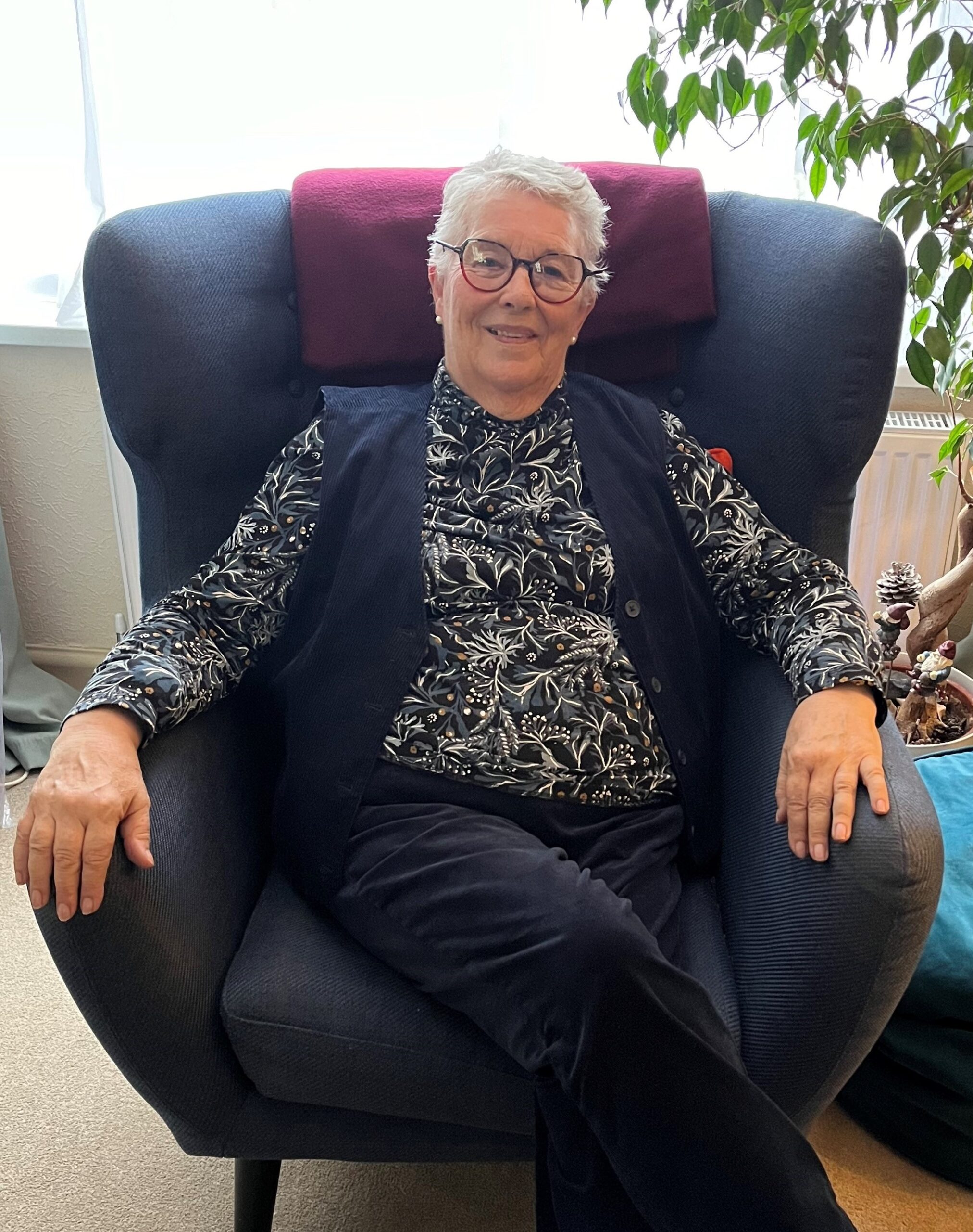 Dawn Horne A woman sits in a blue armchair in front of a window. She has grey hair and is wearing glasses. She is smiling.