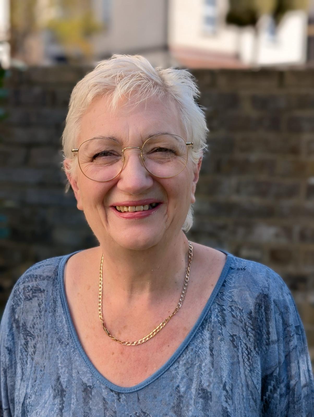 A woman with short grey hair and glasses stands in front of a brick wall, smiling to camera. The light is warm and sunny.