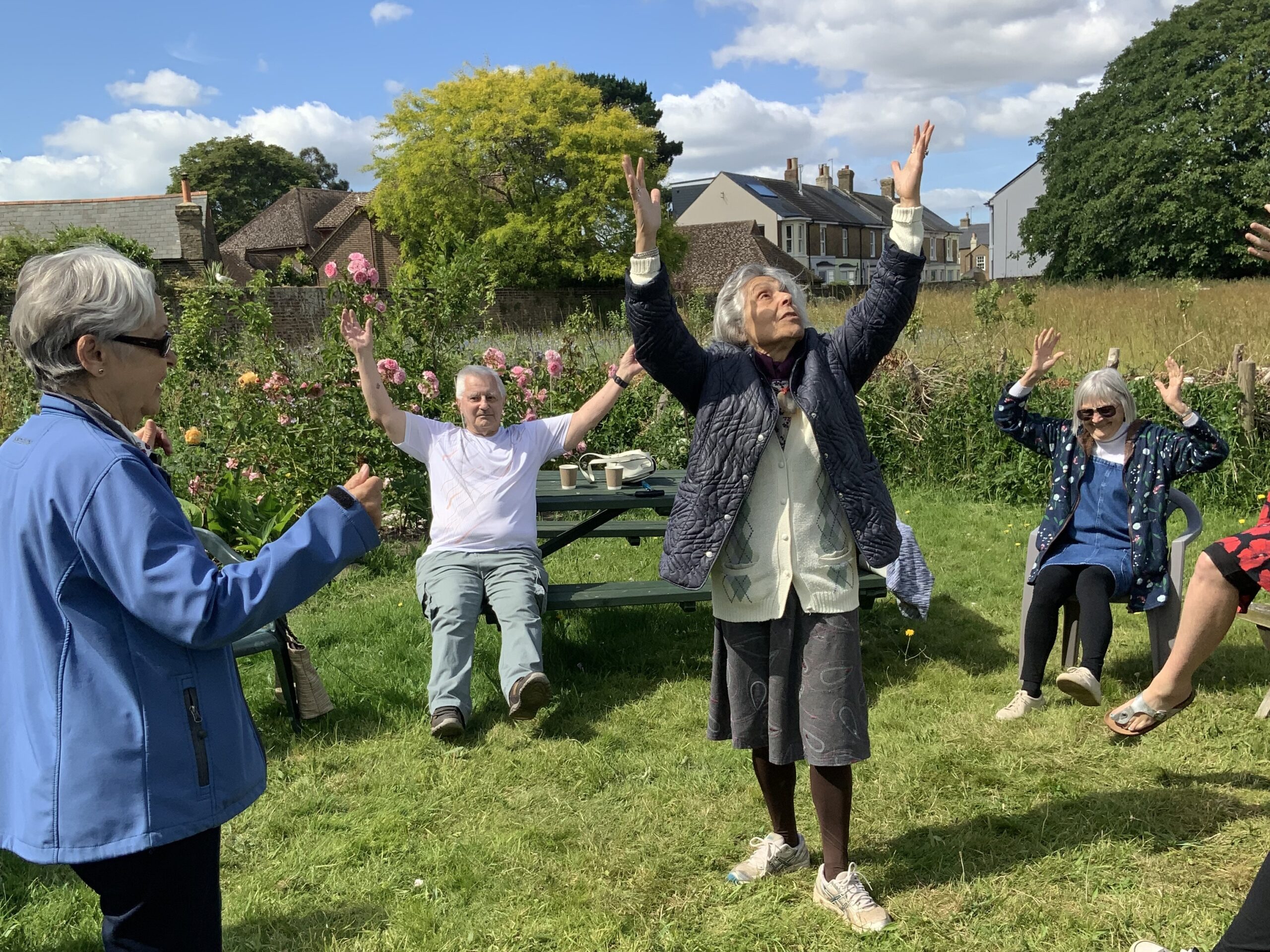 Zest Goes Green 2024 A group of people, some sitting and some on their feet, are dancing in a garden with their arms in the air. It is sunny