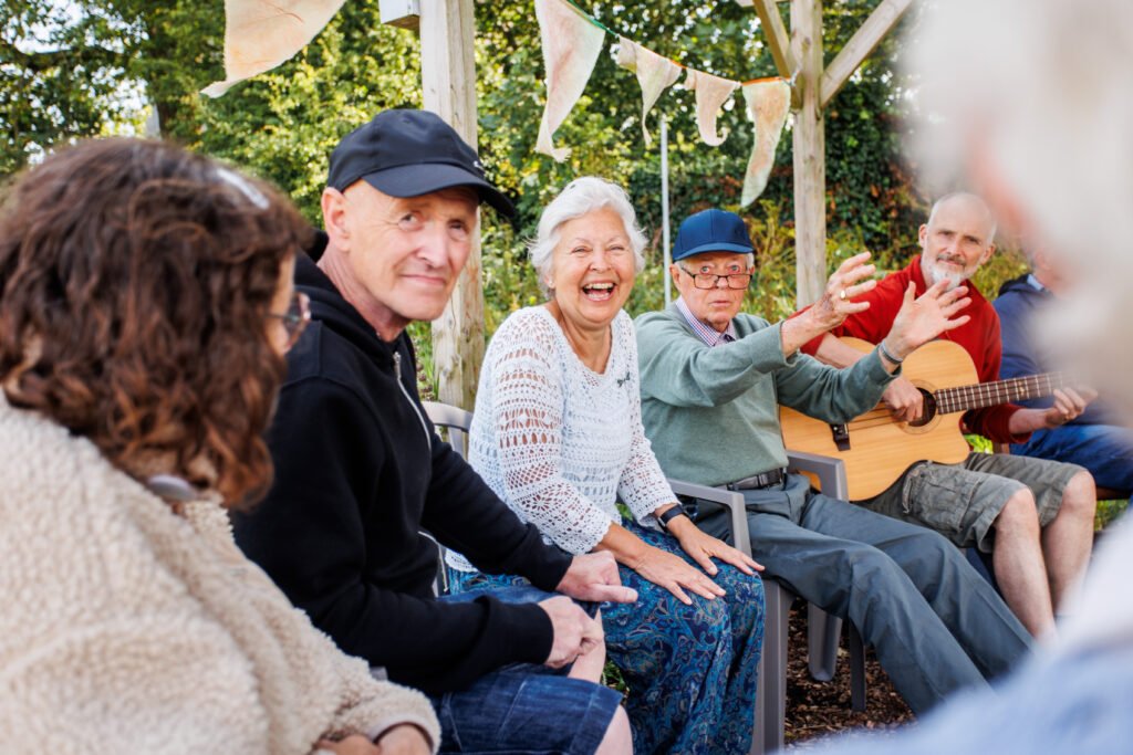 A group of people sit laughing and listening to a man playing an acoustic guitar. They are outside, with bunting above them, looking towards the camera.