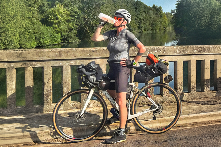 A woman stands on a bridge over a river with her bike, drinking water