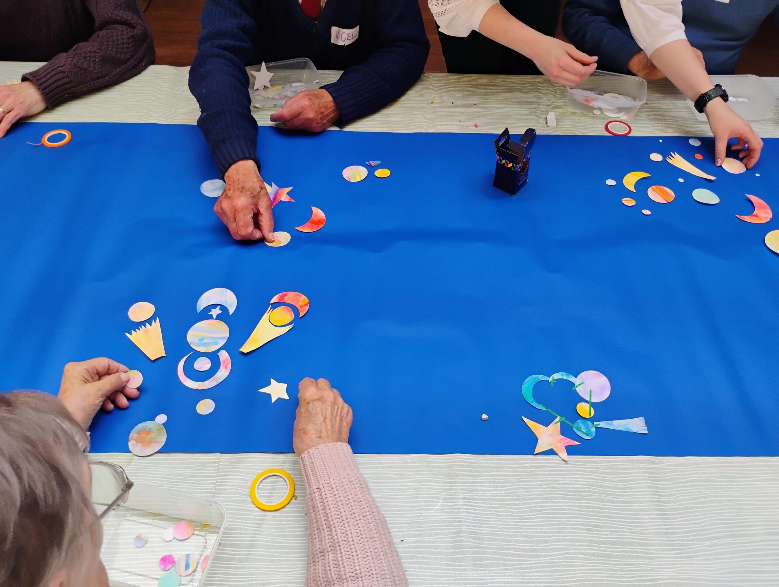 Hands reach across a table covered in blue paper to collage solar systems